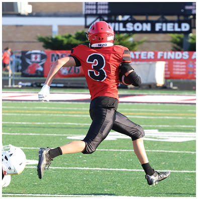 A person wearing a red football jersey with the number three is running on a football field while holding a football. Description generated by AI