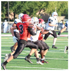People wearing football helmets and uniforms are engaged in a play on a football field with trees in the background. Description generated by AI