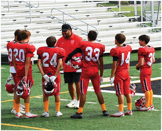 A group of children in red football uniforms with orange socks stand on a field listening to an adult coach wearing a red shirt and black cap. Description generated by AI