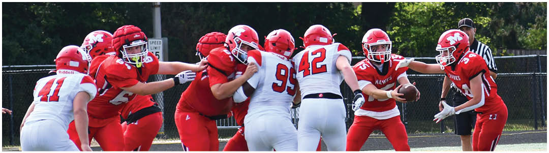 People wearing red and white football uniforms are engaged in a play on a football field with trees and a fence in the background. Description generated by AI