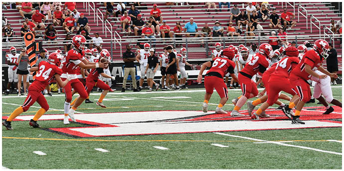 A group of people in red sports uniforms are playing football on a field with spectators seated in bleachers in the background. Description generated by AI
