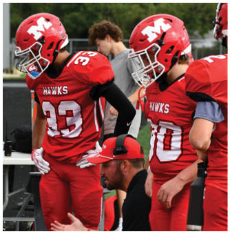 A person wearing a red football uniform with the number 33 and the word “HAWKS" on the jersey is standing next to another person in a gray shirt on a sports field. Description generated by AI
