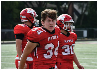Two people wearing red football jerseys with the word “HAWKS" and numbers on the front are standing on a sports field. Description generated by AI