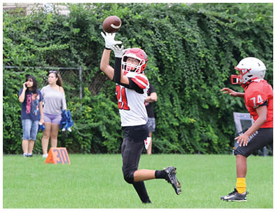 Two children wearing football helmets and uniforms are playing on a grassy field with one child reaching up to catch a ball while the other runs nearby. Description generated by AI