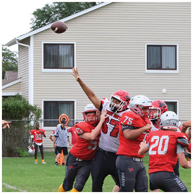 A person in a red football uniform is throwing a football while other players and a referee stand on a grassy field near residential houses. Description generated by AI