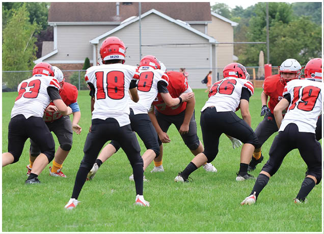 Several people wearing football helmets and uniforms are engaged in a play on a grassy field with houses in the background. Description generated by AI