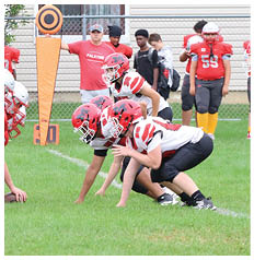 Children in red football uniforms and helmets are positioned on a grassy field preparing for a play while adults stand behind a chain-link fence holding a down marker. Description generated by AI