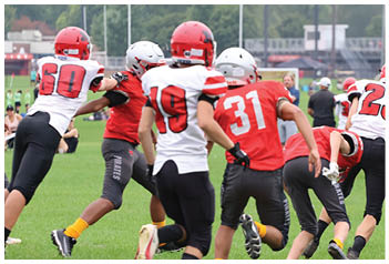 Adults wearing football helmets and uniforms are actively playing on a grassy field with trees and buildings in the background. Description generated by AI