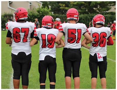 Four people wearing red helmets and white football jerseys with black pants stand side by side on a grassy field with their arms linked behind their backs. Description generated by AI