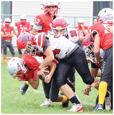 Several people wearing red and white football uniforms and helmets are engaged in a close tackle on a grassy field. Description generated by AI