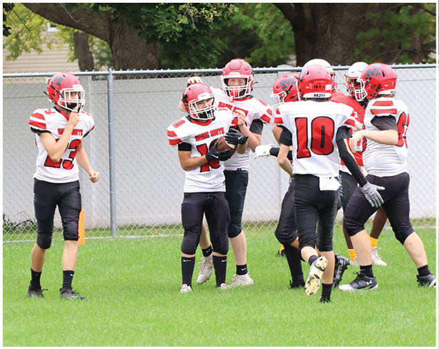 A group of people in red and white football uniforms are standing on a grassy field near a chain-link fence with trees in the background. Description generated by AI