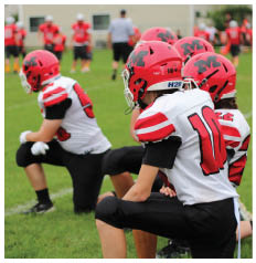 Two people wearing red helmets and white football jerseys with red and black accents are kneeling on a grassy field. Description generated by AI