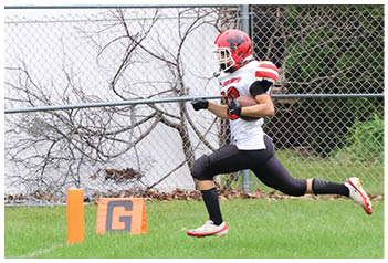 A person wearing a red helmet and black and white sports uniform is running on a grassy field near an orange marker and a chain-link fence with a house in the background. Description generated by AI