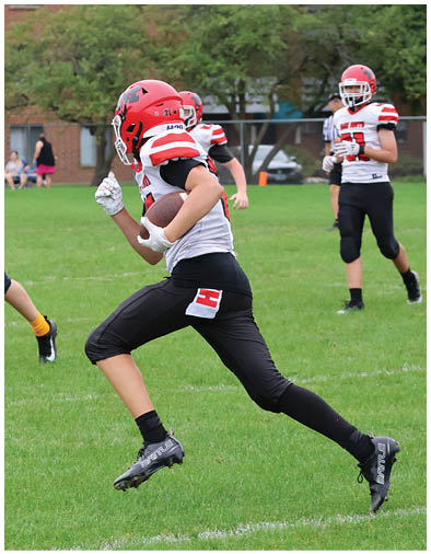 A person wearing a red helmet and black and white football uniform is running on a grassy field while holding a football. Description generated by AI