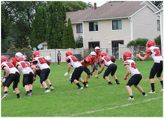 A person in a white and red football uniform is reaching out to catch a football while another person in a red and gray uniform watches in the background on a grassy field. Description generated by AI