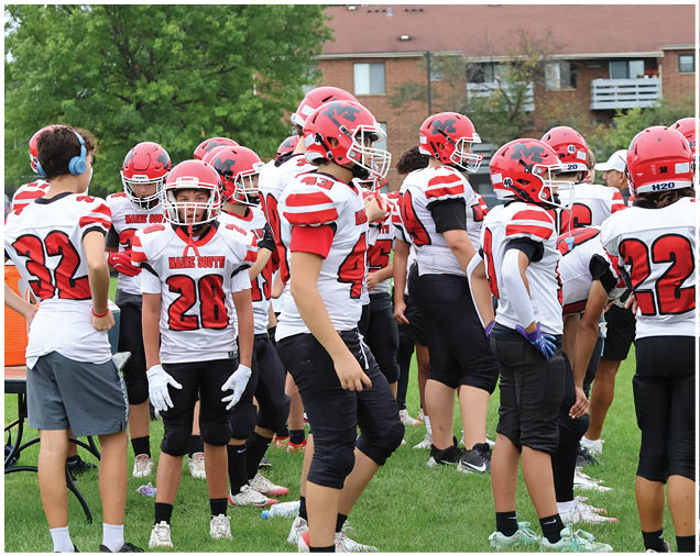 A group of people wearing red and white football uniforms and helmets are gathered on a grassy field near an orange cooler and a table. Description generated by AI