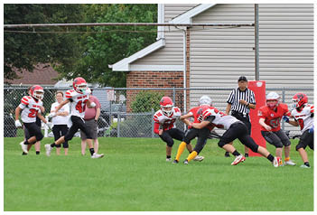 A group of people wearing football helmets and uniforms are playing on a grassy field with a referee standing nearby and a house and trees in the background. Description generated by AI
