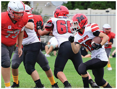 A group of people wearing red helmets and white sports jerseys with red and black accents are gathered on a grassy field, likely preparing for or discussing a football game. Description generated by AI