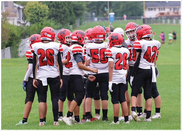 Several people wearing football uniforms and helmets are engaged in a play on a grassy field with a fence and spectators in the background. Description generated by AI