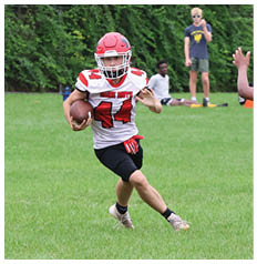 A person wearing a red and white football uniform is running with the ball while another person in a red jersey with the number 74 is preparing to block or tackle on a grassy field with two people standing in the background. Description generated by AI