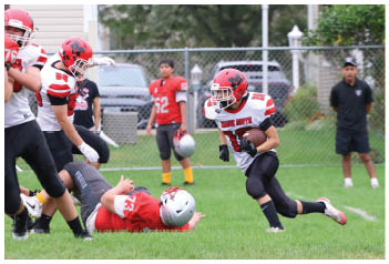 A person in a football uniform is running with the ball while another person lies on the ground and others stand in the background near a chain-link fence. Description generated by AI