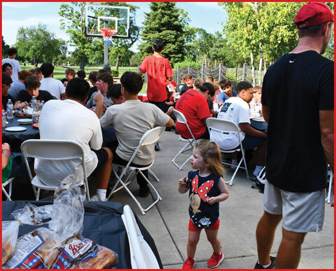 A group of people sitting and eating outdoors near a basketball hoop while a child in a Minnie Mouse outfit walks nearby and an adult in a red cap stands to the side. Description generated by AI