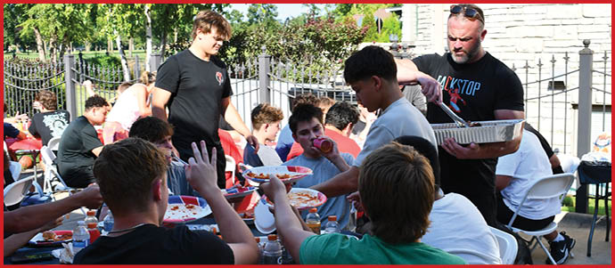 People sitting and standing around a table outdoors sharing food and drinks in a casual social gathering. Description generated by AI