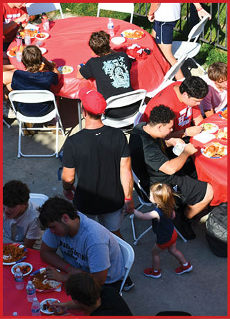 A group of people sitting and standing around red-covered tables outdoors enjoying a meal together on a sunny day. Description generated by AI