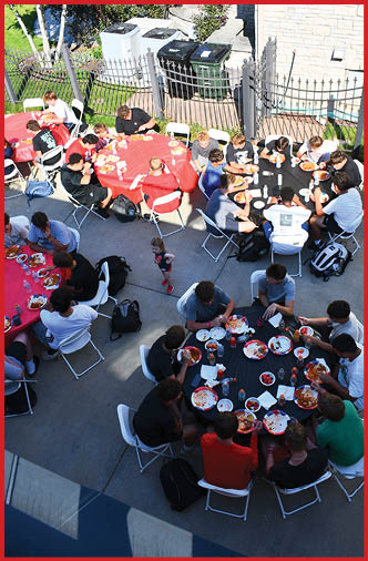 A group of people sitting around a table outdoors eating pizza and drinking bottled water near a metal fence and trash bins. Description generated by AI