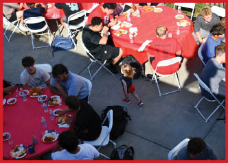 Several people are seated around red tables outdoors eating food while a child in a dark top and red shorts walks nearby on a concrete surface. Description generated by AI