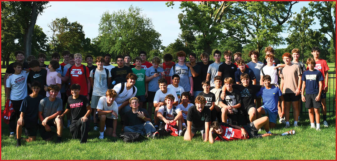 A large group of people wearing casual clothing is gathered outdoors on grass with trees and a clear sky in the background. Description generated by AI