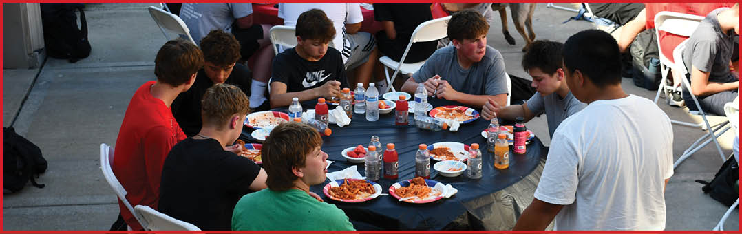 A group of people sitting and standing around a table outdoors, eating pizza and drinking beverages. Description generated by AI