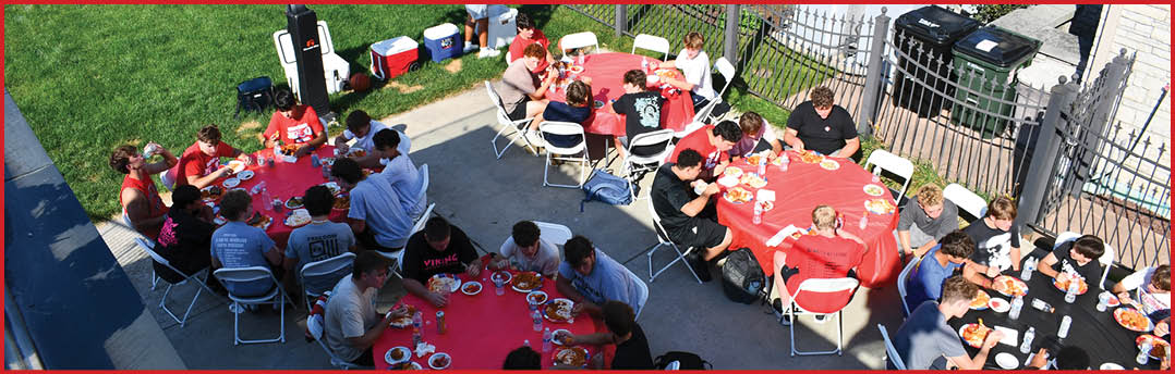 People sitting around red tables outdoors eating and socializing in a casual setting. Description generated by AI