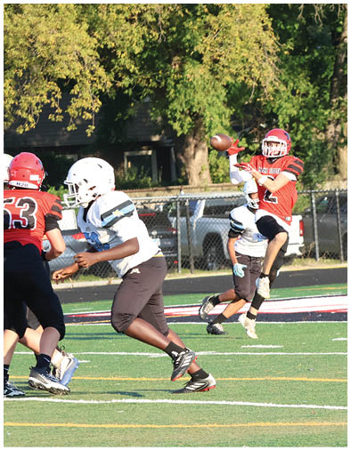 A person in a red sports uniform is jumping to catch a football while another person in a white and blue uniform is running behind on a football field. Description generated by AI