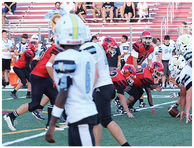 People in football uniforms are lined up on a field preparing for a play with spectators seated in bleachers behind them. Description generated by AI