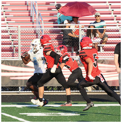 A person in a white football uniform is running with the ball while two people in red football uniforms chase behind on a field with empty bleachers and a few seated people in the background. Description generated by AI