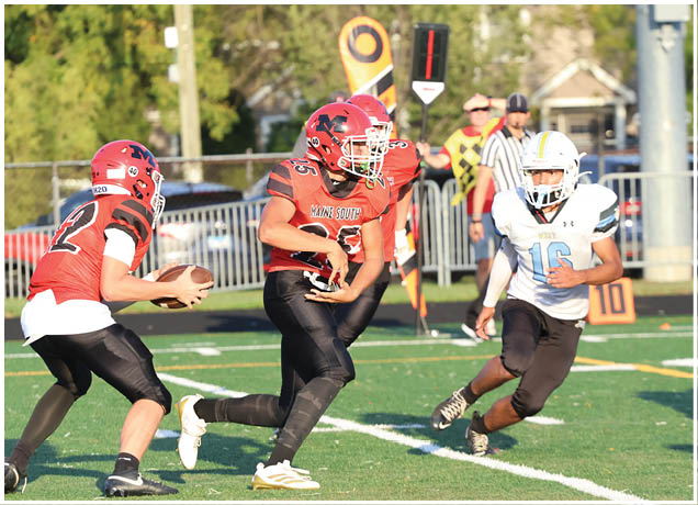 Three people in football uniforms are actively playing on a field, with one person holding a football and another running toward them. Description generated by AI