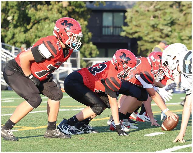Several people wearing red football uniforms and helmets are lined up in a crouched position on a football field, preparing for a play. Description generated by AI