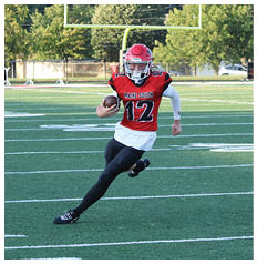 A person wearing a red football uniform with the number 12 is running on a football field holding a football near a goalpost. Description generated by AI