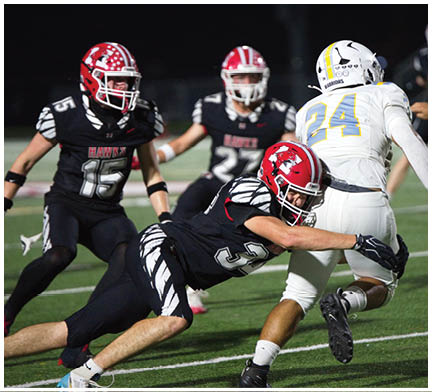 A group of people wearing football uniforms are actively engaged in a game on a field at night, with one person in white running while others in black attempt to tackle. Description generated by AI