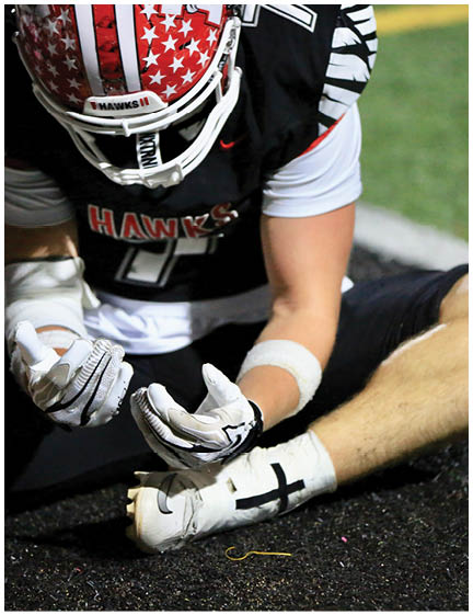 A person wearing a football uniform with “HAWKS" on the jersey is kneeling on the ground with gloved hands near a cleated foot wrapped in white tape with a black cross. Description generated by AI