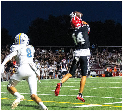 Two people wearing football helmets and uniforms are reaching up to catch a football against a dark sky background. Description generated by AI