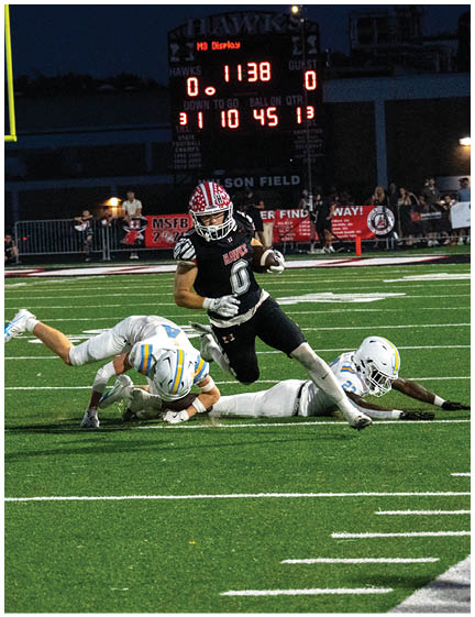 A person wearing a black football uniform with the number 0 is running on a field while two people in white football uniforms are falling or diving near them during a nighttime game. Description generated by AI