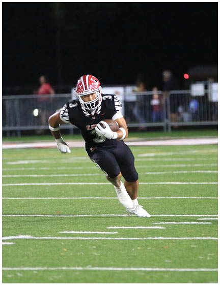 A person wearing a red and white helmet and black sports uniform is running with a football on a lit field at night while a referee and spectators watch in the background. Description generated by AI