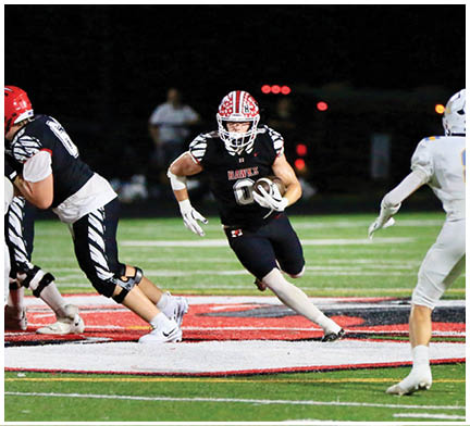 A person in a black football uniform with a red and white helmet is running with the ball while other players in black and white uniforms engage in blocking on a football field at night. Description generated by AI