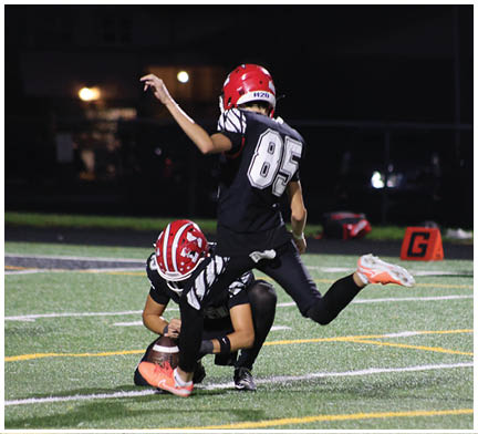 Two people in football uniforms with red helmets are on a field at night, with one holding the ball on the ground and the other preparing to kick it, while a referee watches in the background. Description generated by AI