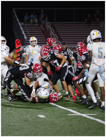 A group of football players in red and black uniforms are tackling a player in a white uniform on a field during a nighttime game. Description generated by AI