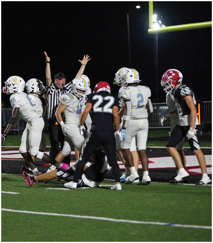 A group of people in football uniforms are gathered on a field with one person raising their arms, possibly a referee signaling a play. Description generated by AI