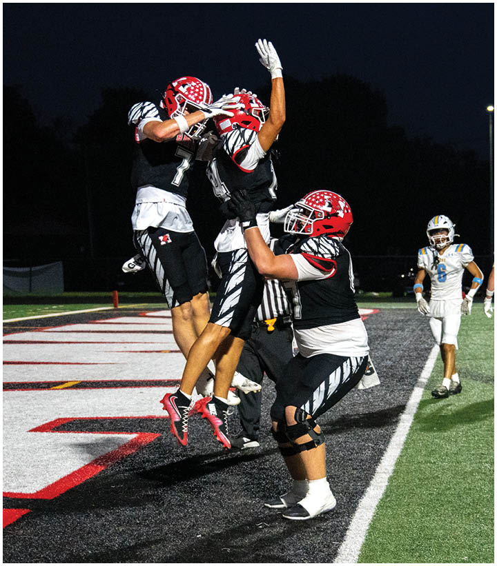 Three football players in black and red uniforms celebrate on a field at night with one player lifting another while a third jumps in the air. Description generated by AI