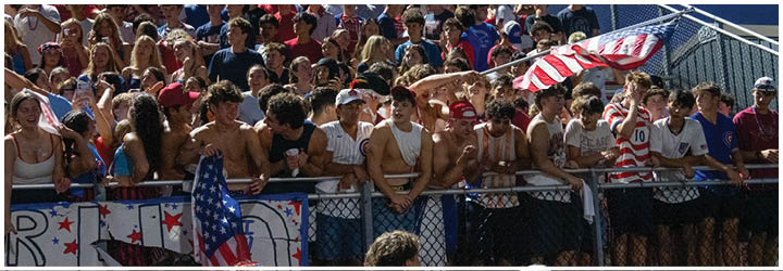 A large crowd of people is seated closely together on bleachers at an outdoor event, with one person waving a red and white striped flag. Description generated by AI